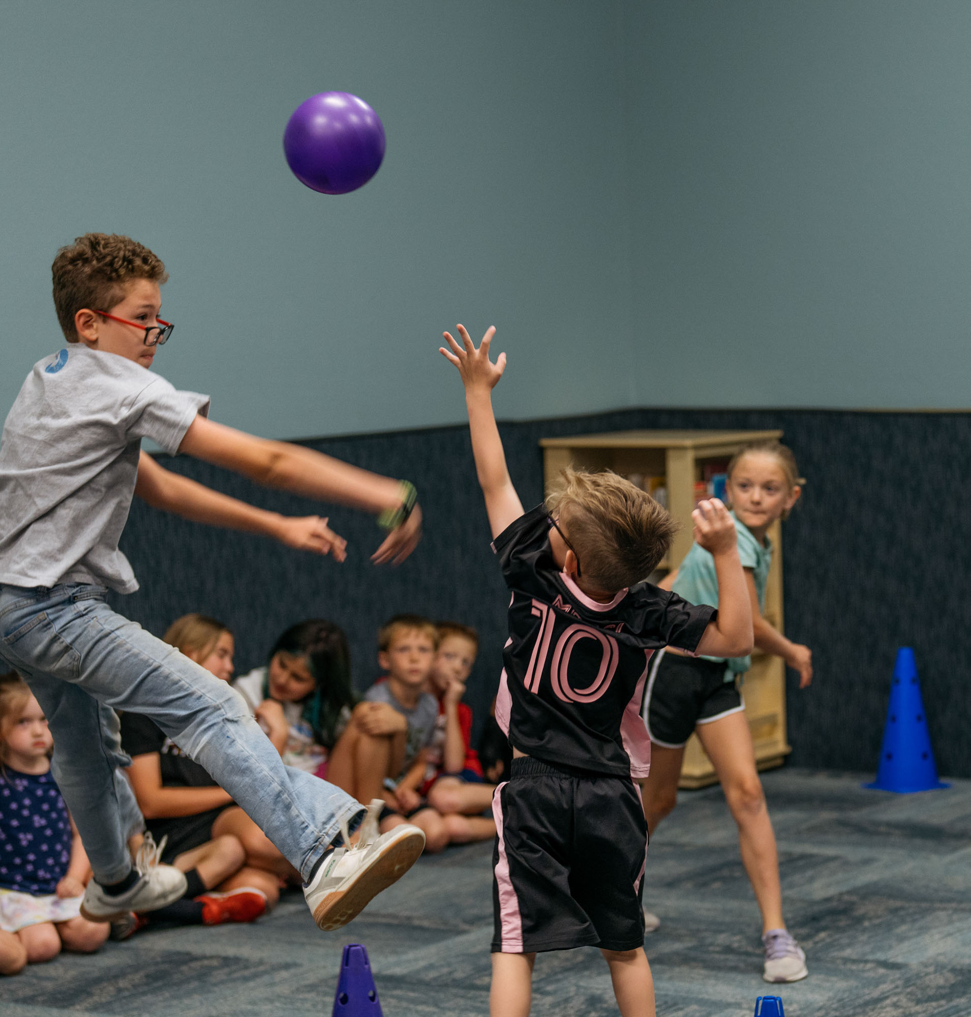 Three children actively play with a purple ball in a room while other kids sit on the floor against the wall watching. Blue cones mark areas on the floor.
