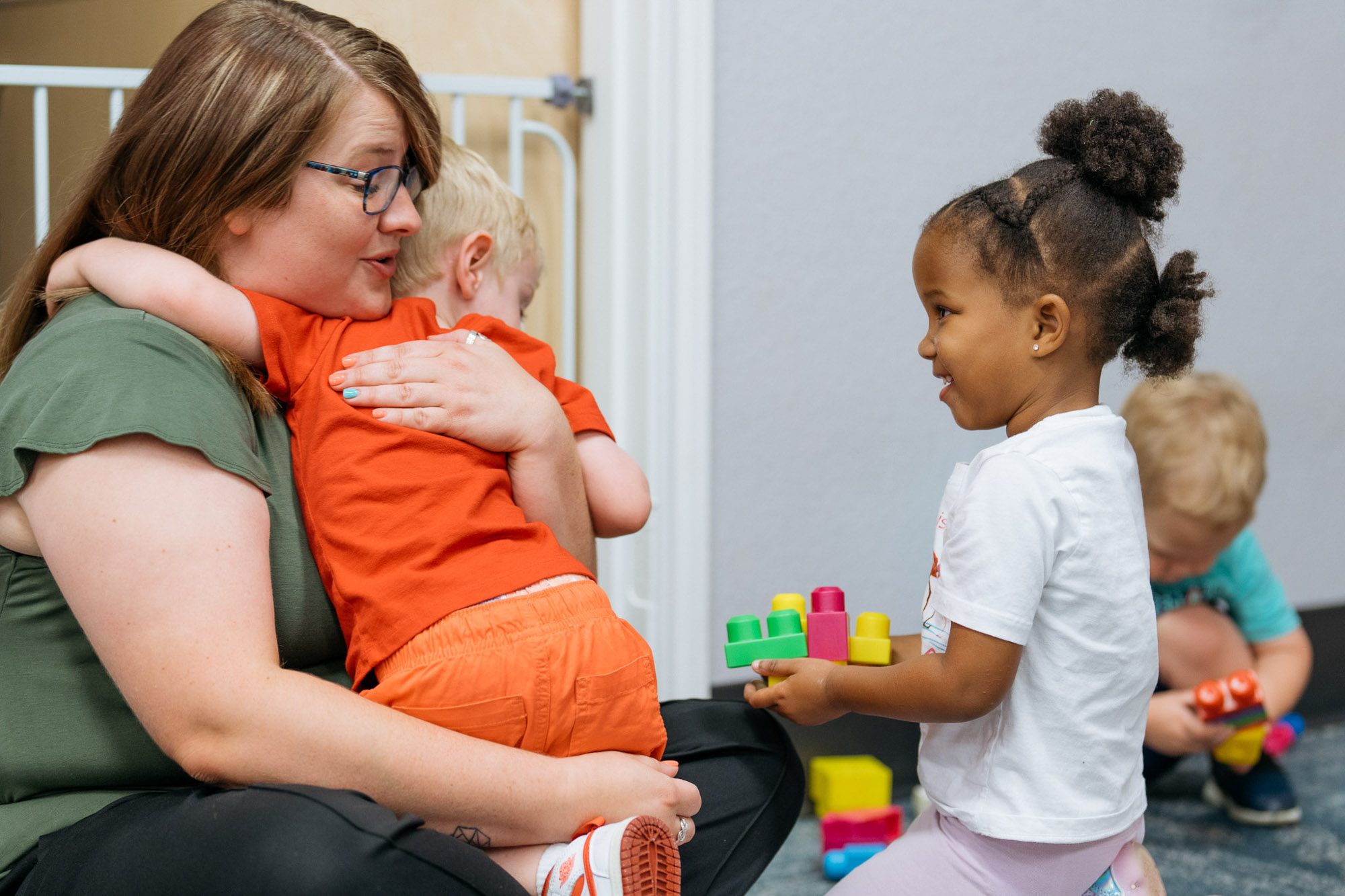 A woman comforts a child in her lap while another child holding toy blocks smiles nearby; a fourth child plays in the background.