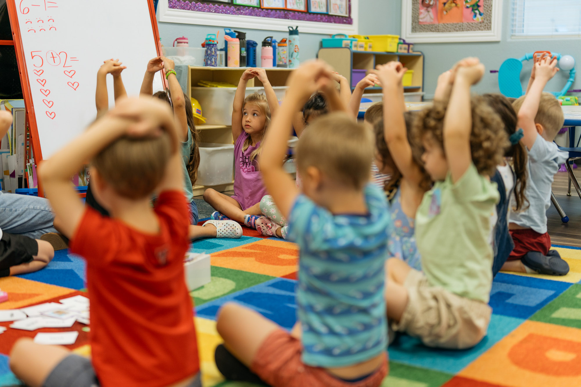 Young children sit in a circle on a colorful classroom rug, raising their arms above their heads, participating in a group activity.