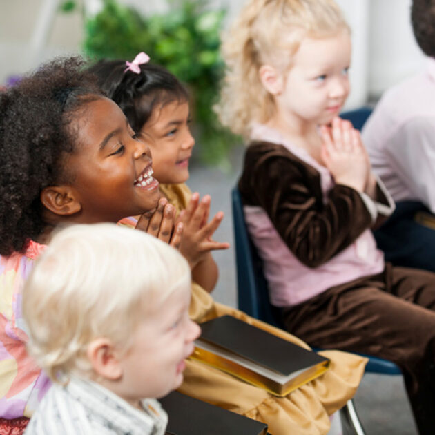 A group of young children sitting in chairs, smiling and clapping their hands, holding books in their laps in a classroom setting.