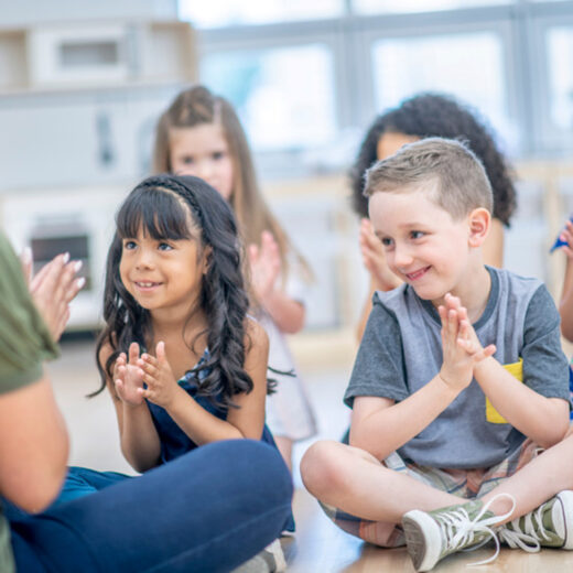 A group of young children sit on the floor in a classroom, clapping and smiling while facing an adult.