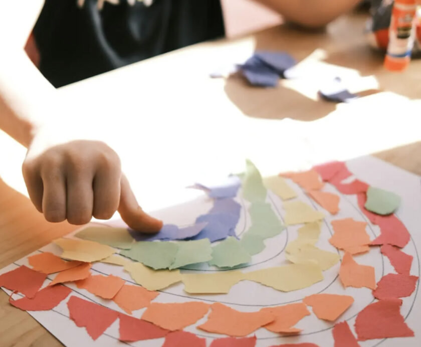 A child’s hand arranges torn colored paper pieces into the shape of a rainbow on a sheet of paper at a table.
