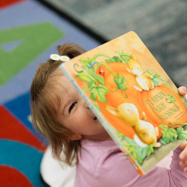 A young child in a pink shirt holds up a colorful picture book, partially covering their face, with a carpet featuring large letters in the background.
