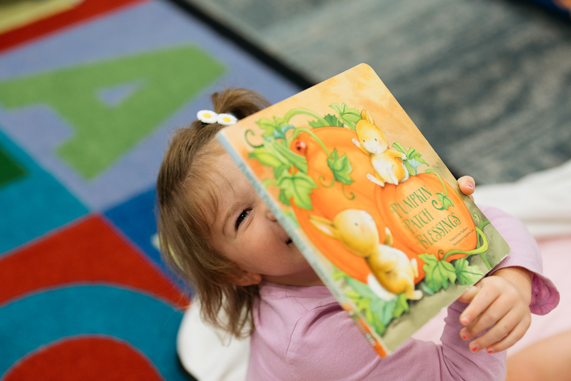 A young child in a pink shirt holds up a colorful picture book, partially covering their face, with a carpet featuring large letters in the background.