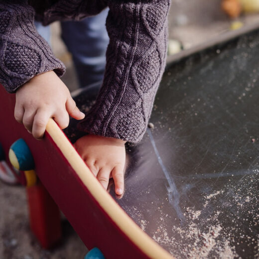 A child wearing a knitted sweater rests their hands on the edge of a sand-covered playground surface.