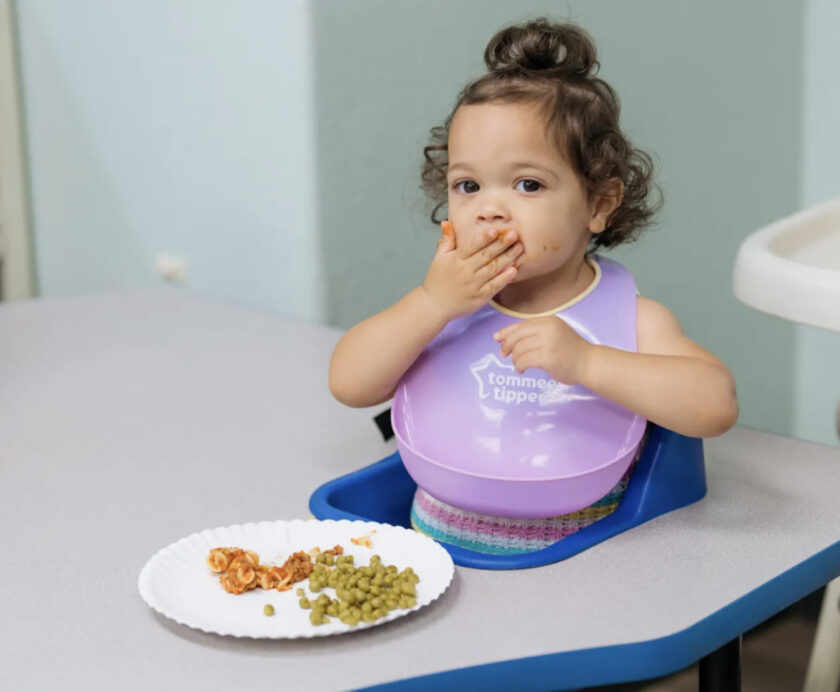 A toddler wearing a purple bib sits at a table, eating pasta and peas from a paper plate, with food on their face and hand.